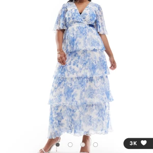 Blommig långklänning med volanger och knytband - Romantisk långklänning i vitt och blått med blommigt mönster, v-ringning och volanger i flera lager. Klänningen har korta ärmar och ett brett knytband i midjan som markerar siluetten. Perfekt för sommarens tillfällen.