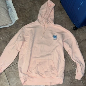 Ljusrosa hoodie från YOURTURN med tryck - Säljer en ljusrosa hoodie från YOURTURN med stor grafisk print på ryggen och mindre tryck på bröstet. Tröjan har huva med snörning och en stor magficka. Materialet är mjukt och skönt, perfekt för chill dagar. Unik design med blått och orange motiv.