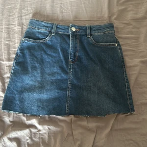 Jeanskjol med rå kant - Säljer en klassisk blå jeanskjol med rå, avklippt kant och femficksmodell. Kjolen har normal passform, bälteshällor och knappstängning framtill. Perfekt till vardags för en avslappnad look! Kjolen är i XS och köpt för väldigt länge sedan! 💌Köparen står för frakten