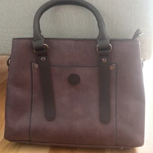 Brun handväska i skinnimitation - Säljer en stilren brun handväska med två handtag och dekorativa remmar framtill. Väskan har en ytterficka med dragkedja på baksidan och detaljer i mörkare brunt. Perfekt för att bära dina viktigaste saker.