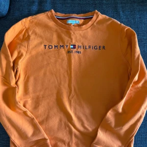 Orange sweatshirt från Tommy Hilfiger - Säljer en orange sweatshirt från Tommy Hilfiger med tryckt logga och text på bröstet. Helt nyskick har aldrig används. Tröjan har rund halsringning och ribbade muddar vid ärmslut och nederkant. Perfekt för en avslappnad stil.