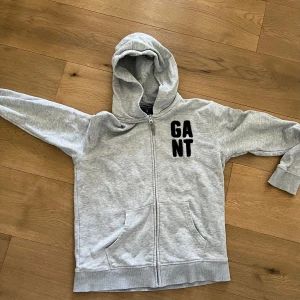 Grå hoodie med dragkedja från GANT - Säljer en ljusgrå hoodie från GANT med svart tryck på bröstet. Tröjan har dragkedja framtill, huva och två fickor. Perfekt för en avslappnad stil och enkel att matcha med det mesta.