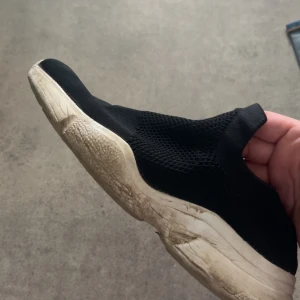 Svarta slip-on sneakers med vit sula - Svarta slip-on sneakers med ovandel i mesh och elastiskt material. Skon har en tjock vit sula och en sportig, minimalistisk design utan snörning. Perfekt för en avslappnad och bekväm stil. De har blivit väl använda av en kvinna.