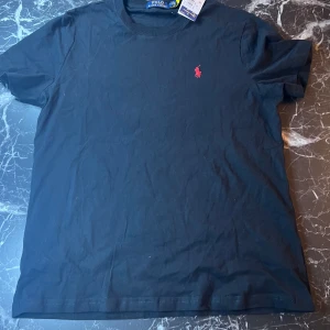 Svart t-shirt från Polo Ralph Lauren - Säljer en svart t-shirt från Polo Ralph Lauren med klassisk röd broderad logga på bröstet. T-shirten har rund halsringning och är kortärmad. Perfekt för en stilren och enkel look.