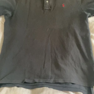Svart pikétröja från Ralph Lauren - Klassisk svart pikétröja från Ralph Lauren med röd broderad logga på bröstet. Tröjan har krage, två knappar framtill och är tillverkad i bomull. Perfekt för en clean och stilren look.