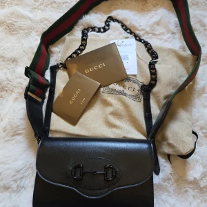 Gucci Horsebit 1955 väska - Säljer en svart axelväska från Gucci i äkta skinn med klassisk hästbitsdetalj på framsidan. Väskan har bred axelrem med gröna och röda ränder samt en kedja och levereras med dustbag och äkthetskort. Perfekt för dig som gillar lyxiga accessoarer.