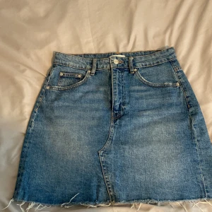 Jeanskjol från Gina Tricot  - Klassisk blå jeanskjol från Gina Tricot med femficksdesign och råa kanter nertill. Kjolen har normal passform och är i ett tåligt denimtyg. Perfekt för dig som gillar en enkel och tidlös stil.