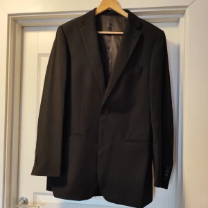 Svart kavaj från Dressmann - Klassisk svart kavaj från Dressmann i storlek 52.. Kavajen har två knappar framtill, fickor med lock och en bröstficka. Perfekt för formella tillfällen och enkel att matcha med skjorta och slips. Använd en gång