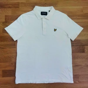 Vit pikétröja från Lyle & Scott - Klassisk vit pikétröja från Lyle & Scott med krage och knäppning framtill. Tröjan har korta ärmar och den ikoniska gula loggan broderad på bröstet. Perfekt för en stilren och sportig look.
