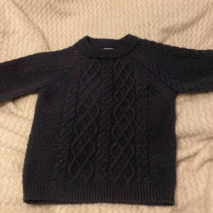 Mörkblå stickad tröja från 157 - Mysig mörkblå stickad tröja från 157 med klassiskt flätmönster på framsidan och strukturstickning på baksidan och ärmarna. Tröjan har rund halsringning och långa ärmar med ribbade muddar.