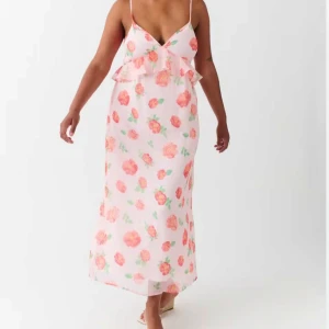 Blommig långklänning med volang - Söt långklänning med rosa och röda blommor på ljus botten. Klänningen har smala axelband, v-ringning och volangdetalj framtill. Perfekt för sommarens alla tillfällen.