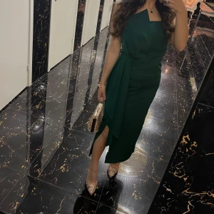 Mörkgrön klänning  - Säljer en elegant mörkgrön klänning med omlottdesign. Klänningen har en asymmetrisk nederdel med liten slits. Perfekt för dig som vill sticka ut med stil.