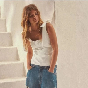 INTRESSEKOLL Vit linnetopp med rosett - Säljer en stilren vit linnetopp med breda axelband och en söt rosettdetalj på axeln. Toppen har en rak passform och är perfekt för varma dagar.