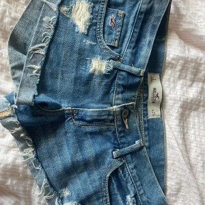 Blå jeansshorts med spets från Hollister - Säljer ett par blå jeansshorts från Hollister med fransade kanter och slitningar. Shortsen har spetsdetaljer på insidan av benen och klassisk femficksdesign. Perfekta för varma dagar och har låg midja. Storlek W25.