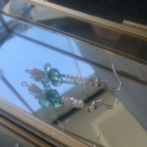 Handgjorda örhängen med pärlor i blått glas - Unika örhängen med vita pärlor och glittrande blågröna glaspärlor i olika former. Droppformad vit pärla längst ner och små runda pärlor upptill. Hängande modell med silvriga krokar, perfekt för dig som gillar skimmer och detaljer.