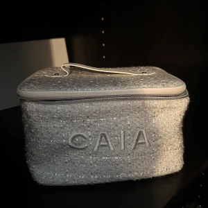 Grå necessär från CAIA med dragkedja - Säljer en snygg och rymlig necessär från CAIA i ljusgrått bouclé-liknande tyg med broderad logga framtill. Väskan har ett praktiskt handtag upptill och stängs med dragkedja. Perfekt för smink eller hudvård när du är på språng.