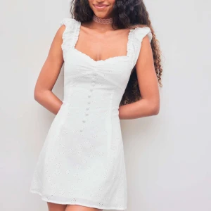 Vit broderad kortklänning med volang - Superfin vit kortklänning med broderade blommor, volangdetaljer vid axlarna och knappar framtill. Perfekt klänning inför studenten eller midsommar. 