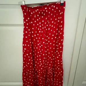 Röd prickig långkjol med volanger - Superfin röd långkjol med vita prickar och volangdetaljer. Kjolen har en avslappnad passform och resår i midjan för extra komfort. Perfekt för dig som vill sticka ut med en färgglad och lekfull stil.