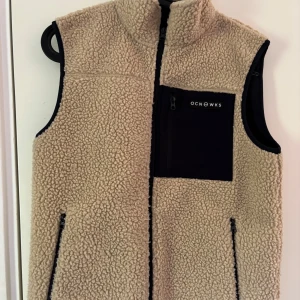 Beige fleeceväst från OCNWKS - Mysig beige fleeceväst från OCNWKS med svart bröstficka och dragkedja framtill. Västen har hög krage och svarta detaljer vid fickor och ärmkanter. Perfekt lager för kyligare dagar.