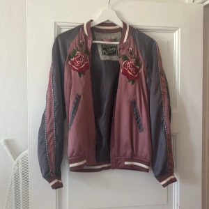Rosa och grå bomberjacka med broderade rosor - Säljer en snygg bomberjacka i rosa och grått från Replay, Jackan har broderade röda rosor på bröstet, randiga muddar och dragkedjefickor. Perfekt för dig som vill sticka ut lite extra!
