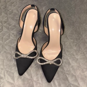 Svarta pumps med rosett och klack - Säljer ett par eleganta svarta pumps med spetsig tå och glittrig rosettdetalj framtill. Skorna har öppen häl och en unik, böjd klack som ger en trendig look. Perfekta för dig som vill sticka ut med stilrena detaljer.