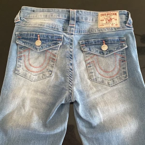 Ljusblå bootcut jeans från True Religion - Ljusblå jeans från True Religion med bootcut passform och mid rise. Fina rosa detaljer. Köptes på Plick men tyvärr lite för små för mig. Midjemått: 38 cm. Innerbenslängd: 81 cm. 