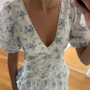 Vit blommig klänning  - Superfin vit klänning med blått blommönster och v-ringning. Klänningen har volanger i kjolen och spetsdetaljer längs med sömmarna. Hör av er om ni har frågor eller vill ha fler bilder, priset går att diskutera💞