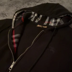 Svart hoodie med dragkedja från Burberry - Svart hoodie från Burberry med dragkedja och klassiskt rutigt foder i huvan. Snygg detalj med märkets logga och mönster i svart, vitt och rött. Perfekt för en stilren och avslappnad look. (9/10 skick✅) Underbar till sommaren och besviker aldrig 🙅 . Skriv gärna ifall du har några frågor!!!