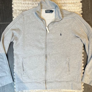 Ralph Lauren Grå Full Zip - Klassisk hoodie med dragkedja från Ralph Lauren i grått. Mjuk bomullskvalitet och en tidlös look. Perfekt basplagg i garderoben. 🔹 Storlek: M 🔹 Färg: Grå 🔹 Skick: Mycket fint skick   