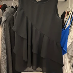 Svart volangtopp - Säljer en svart blus från H&M med flera lager volanger som ger en snygg och flowy look. Blusen är ärmlös och har en rund halsringning. 