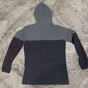 Grå och svart vindjacka från Under Armour - Säljer en snygg tvåfärgad vindjacka från Under Armour i grått och svart. Jackan har huva, dragkedja framtill och långa ärmar. Perfekt för träning eller utomhusaktiviteter. Logga på bröstet och justerbar huva.