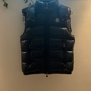 Svart dunväst från Moncler - Säljer en svart, glansig dunväst från Moncler med hög krage och dragkedja framtill. Västen har två fickor med dragkedja och ikonisk Moncler-logga på bröstet. Insidan har ett serietidningsmönster och märkets etikett. Fick den i present så har tyvärr ingen möjlighet att visa er kvitto eller liknande! Därför så går jag ner lite i pris för att visa lite respekt! Använt fåtal gånger men det märks inte. Har använt västen utan luva så luvan är som ny. Pris kan diskuteras. Hör av er om ni är intresserad