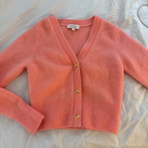 Rosa kofta från & Other Stories - Superfin rosa kofta från & Other Stories med v-ringning och unika guldfärgade delfinknappar. Koftan är croppad, har långa ärmar och är stickad i ett mjukt material som känns lyxigt. Perfekt för att lyfta din outfit med en söt detalj. Kan även gå ner i pris vid snabb affär