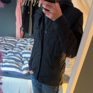 marinblå j.lindeberg field jacka - Säljer en marinblå vindjacka från Jlindeberg med snygga detaljer som bröstfickor, dragkedja och knappar framtill. Jackan har hög krage och flera praktiska fickor. Perfekt för dig som vill ha en stilren och funktionell jacka till vardags. obs en knapp har gått av men den medföljer jättelätt att sy på därav billigare, inköpt för 3000
