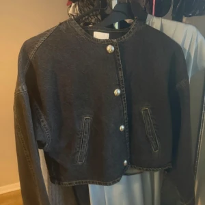 Svart croppad jeansjacka från H&M - Svart croppad jeansjacka från H&M med rund halsringning och coola pärlknappar framtill. Jackan har två fickor framtill och markerade sömmar som ger en snygg vintagekänsla. Perfekt för dig som vill ha en stilren men edgy look.