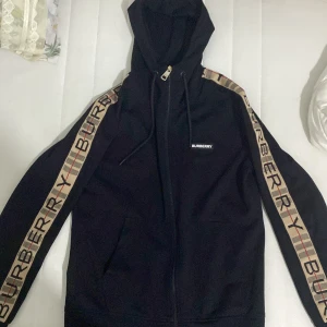 Svart zip hoodie från Burberry - Säljer en snygg svart hoodie från Burberry med dragkedja och huva. Den har märkets logga på bröstet och ikoniska ränder längs ärmarna. Perfekt för en stilren look.Har använt den jätte lite 