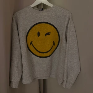 Grå sweatshirt med smiley - Säljer en grå sweatshirt med en stor gul smiley på framsidan från Zara. Tröjan har långa ärmar och en rund halsringning. 