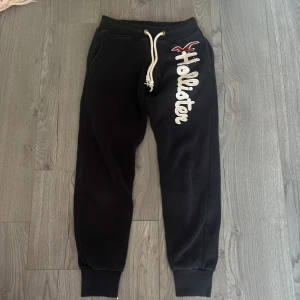 Svarta mjukisbyxor från Hollister - Säljer ett par svarta mjukisbyxor från Hollister med vit snörning i midjan och ett stort broderat Hollister-logotyp på benet. Byxorna har ribbade muddar vid bensluten och är perfekta för en avslappnad stil. Går att diskutera pris.