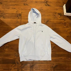 Ljusblå zip hoodie från Polo Ralph Lauren - Säljer en stilren ljusblå hoodie från Polo Ralph Lauren med dragkedja och broderad logga på bröstet. Perfekt för en avslappnad look. Tyvärr är dragkedjan trasig men tröjan är i jätte fint skick annars. Om man vill fixa kedjan är det fritt fram, men där av det låga priset.