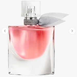 Lancome parfym - Elegant parfymflaska med en mjukt rosa vätska. Flaskan har en fyrkantig form med en klar glaskork. Runt halsen finns en glittrande silverdetalj och en dekorativ silverfärgad rosett. Perfekt för den som älskar en lyxig och feminin doftupplevelse.