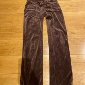 Bruna mjukisbyxor från Juicy Couture - Säljer ett par bruna mjukisbyxor från Juicy Couture i storlek XS. Byxorna har en bekväm passform med fickor fram och bak samt en dragsko i midjan. Perfekta för en avslappnad stil.