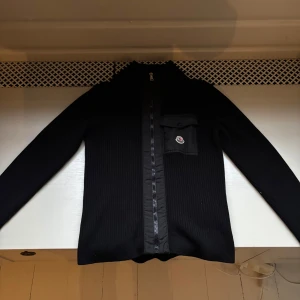 Svart ribbad tröja från Moncler - Snygg svart ribbad tröja från Moncler med dragkedja och en liten ficka på bröstet. Perfekt för kyligare dagar med sin stilrena design och långa ärmar. Tröjan har en diskret logga på fickan.