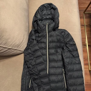 Svart dunjacka från Michael Kors - Snygg svart dunjacka från Michael Kors med huva och dragkedja. Jackan har en quiltad design och långa ärmar. Perfekt för kyligare dagar.