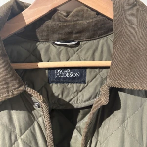 Grön quiltad jacka från Oscar Jacobson - Säljer en stilren grön quiltad jacka från Oscar Jacobson. Jackan har en brun manchesterkrage och knäppning framtill. Två stora fickor med lock på framsidan ger extra funktionalitet. Perfekt för kyligare dagar!