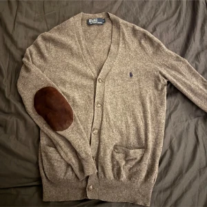 Grå kofta från Polo Ralph Lauren - Säljer en stilren grå kofta från Polo Ralph Lauren med bruna armbågslappar. Koftan har knappar framtill och två fickor. Perfekt för en klassisk look.