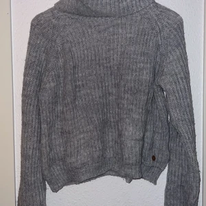 Grå stickad polotröja från Ü - Säljer en mysig grå stickad polotröja från Ü. Tröjan har en hög krage och långa ärmar, perfekt för kyliga dagar. Den ribbade strukturen ger en stilren look. Passar utmärkt för både vardag och avslappnade tillfällen.
