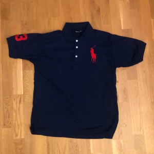 Mörkblå pikétröja från Ralph Lauren - Säljer en klassisk mörkblå pikétröja från Ralph Lauren med röd logga på bröstet och nummerdetalj på ärmen. Tröjan har korta ärmar och tre knappar framtill. Perfekt för en stilren look.