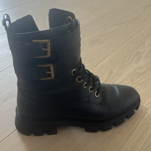 Svarta boots med spännen från Michael Kors - Snygga svarta boots i skinn från Michael Kors med robust sula och guldfärgade detaljer. Skorna har snörning framtill och två dekorativa spännen på skaftet. Perfekta för en stilren look.