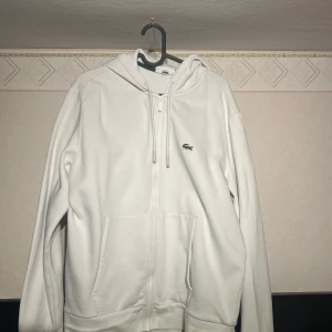 Vit hoodie från Lacoste - Säljer en stilren vit hoodie från Lacoste med dragkedja och klassisk krokodil-logga på bröstet. Perfekt för en avslappnad stil. Den har en huva med dragsko och praktiska fickor framtill. Storlek XL men passar L