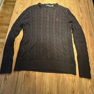 Mörkblå tröja från Polo Ralph Lauren - Säljer en stilren mörblå kabelstickad tröja från Polo Ralph Lauren. Tröjan har en klassisk design med ett rött broderat logotypmärke på bröstet. Perfekt för kyligare dagar med sin mjuka och bekväma passform. Den är lite töjd vid nacken och höften.